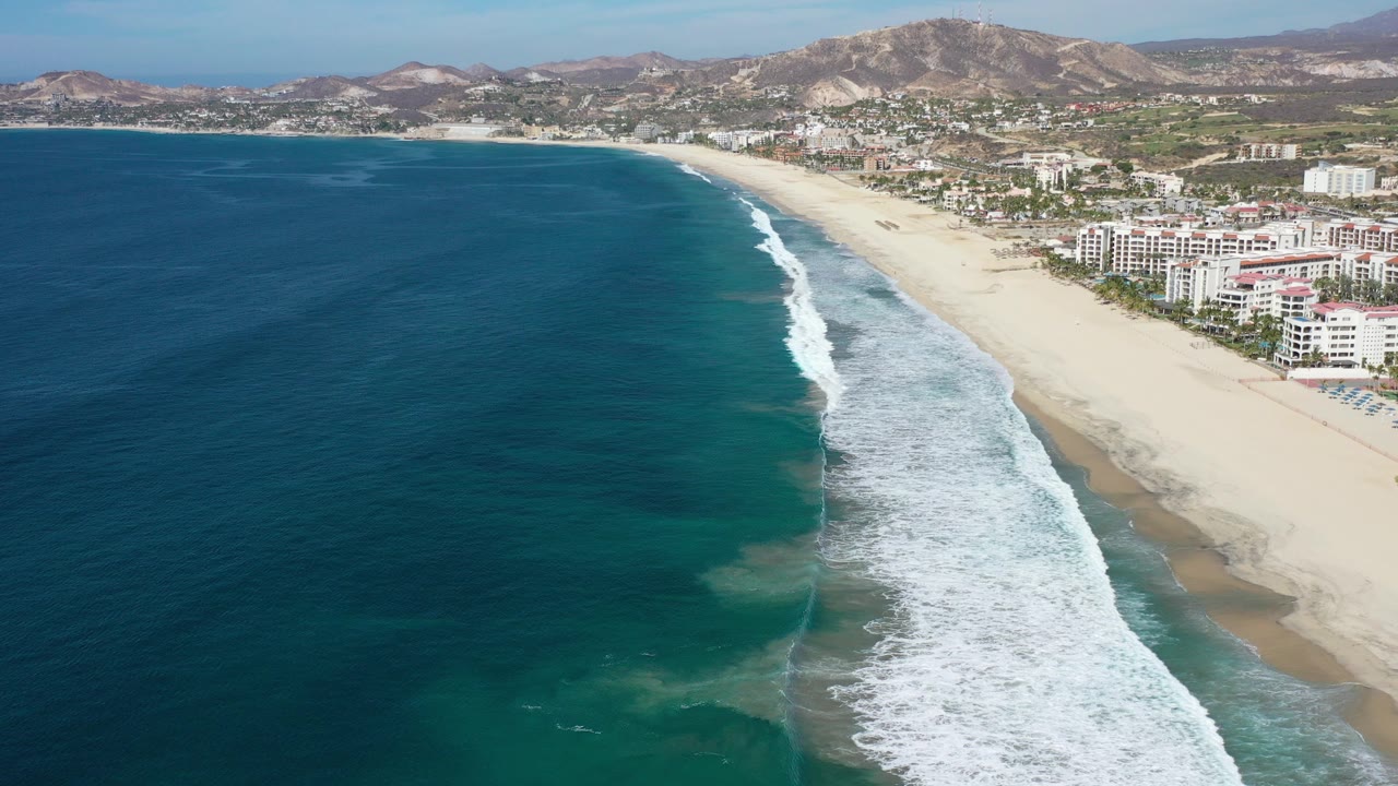Aerial view over Los Cabos, Mexico showing deep blue ocean at left, a white surf line on sandy beach, waterfront buildings, inland hills, and no visible people.