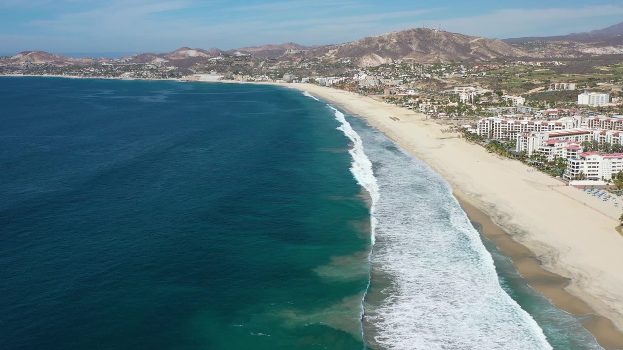 Wide aerial coastline with dark ocean, bright surf, beach, town, and hills.