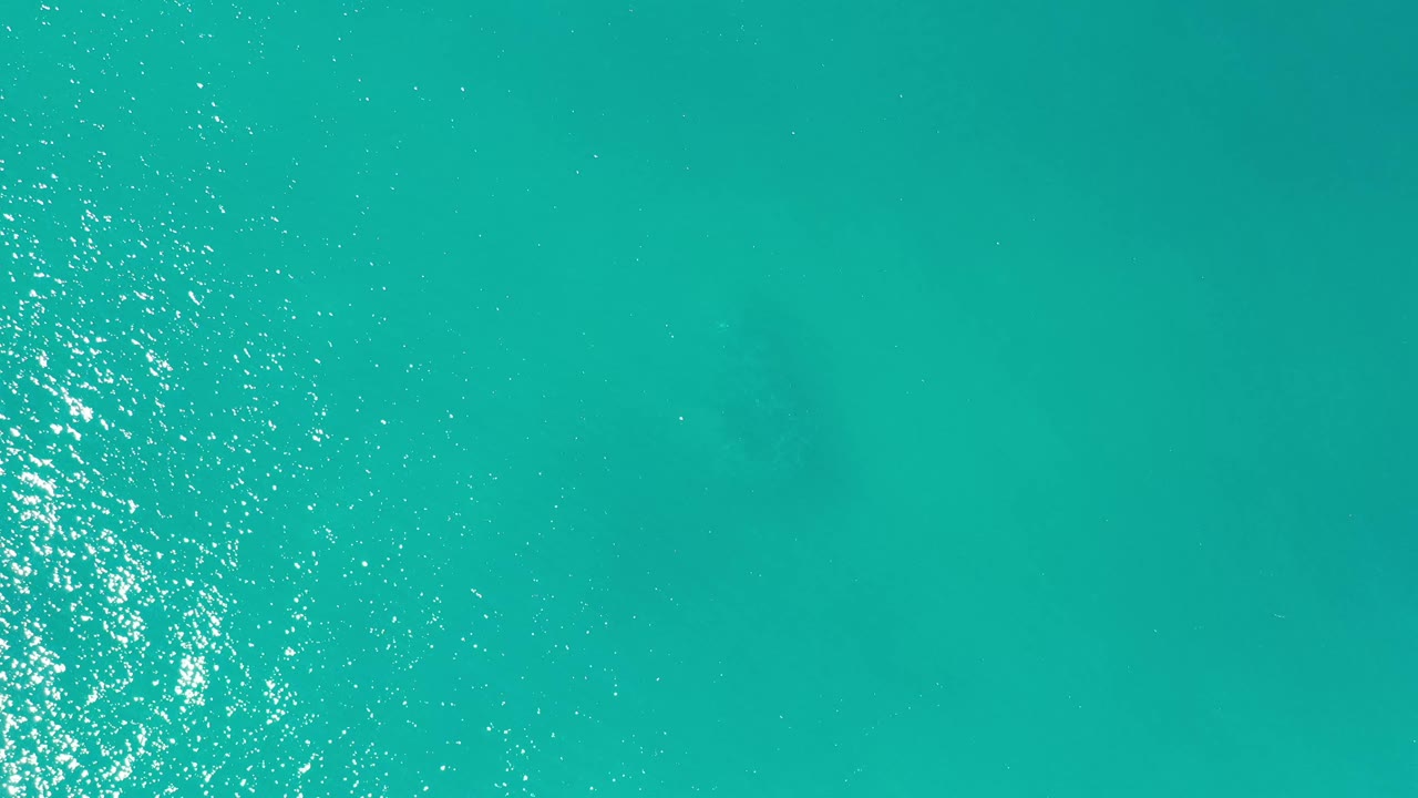 Aerial ocean frame from Los Cabos, Mexico, showing turquoise water, a central vertical strip of sun reflection, faint darker patches below the surface, and no visible people.