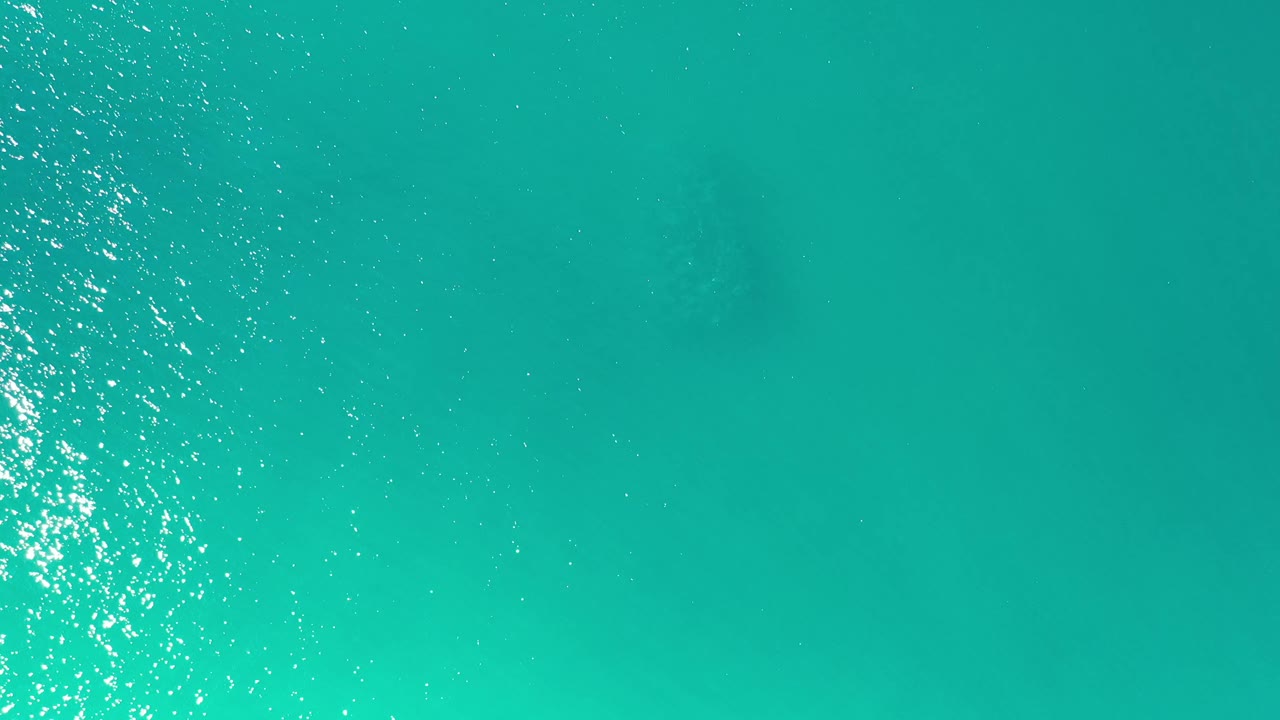 Top-down Los Cabos water frame with turquoise color, faint darker patches below the surface, a centered band of sun reflection, and no visible people.