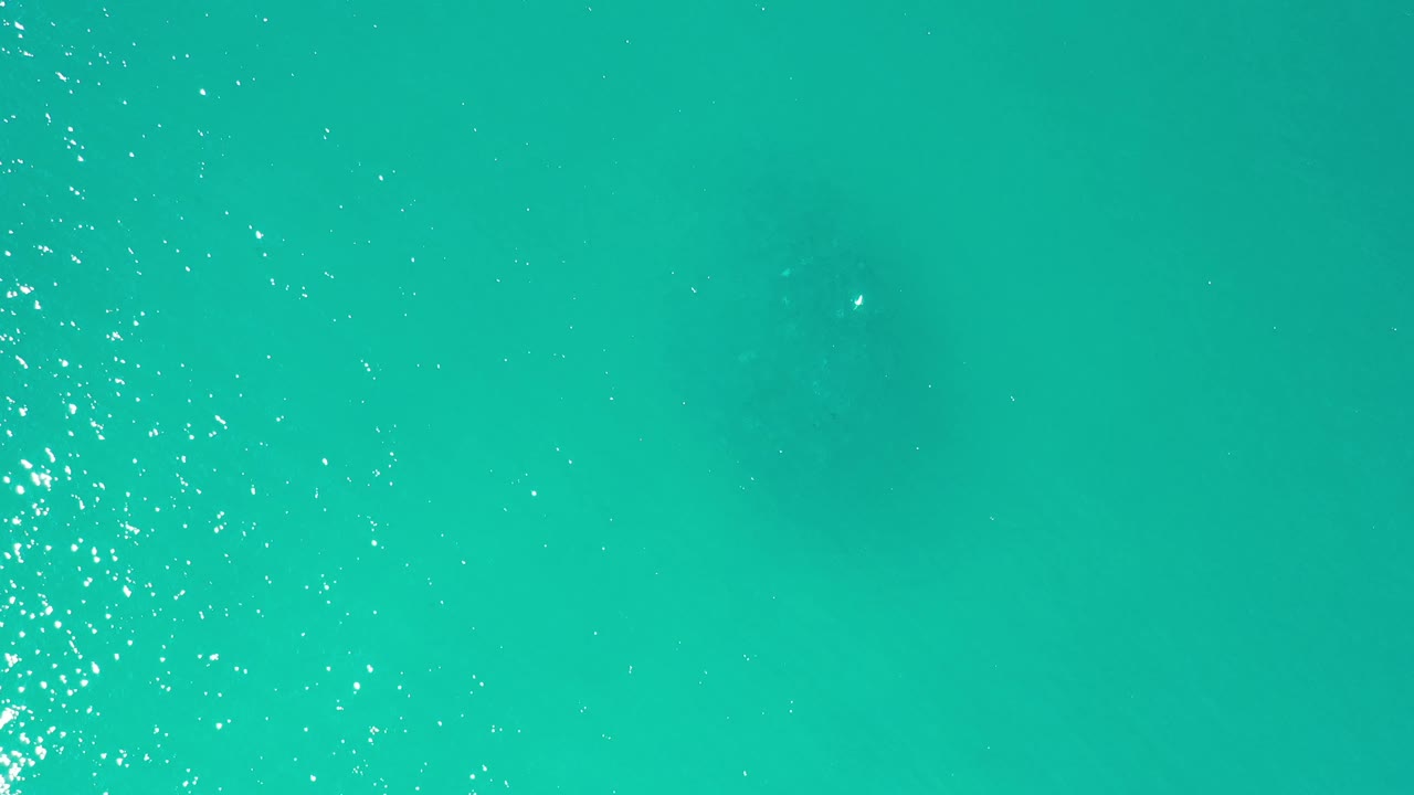 Aerial frame over clear green-blue water in Los Cabos, Mexico, with a bright vertical band of surface sparkle and no visible people.