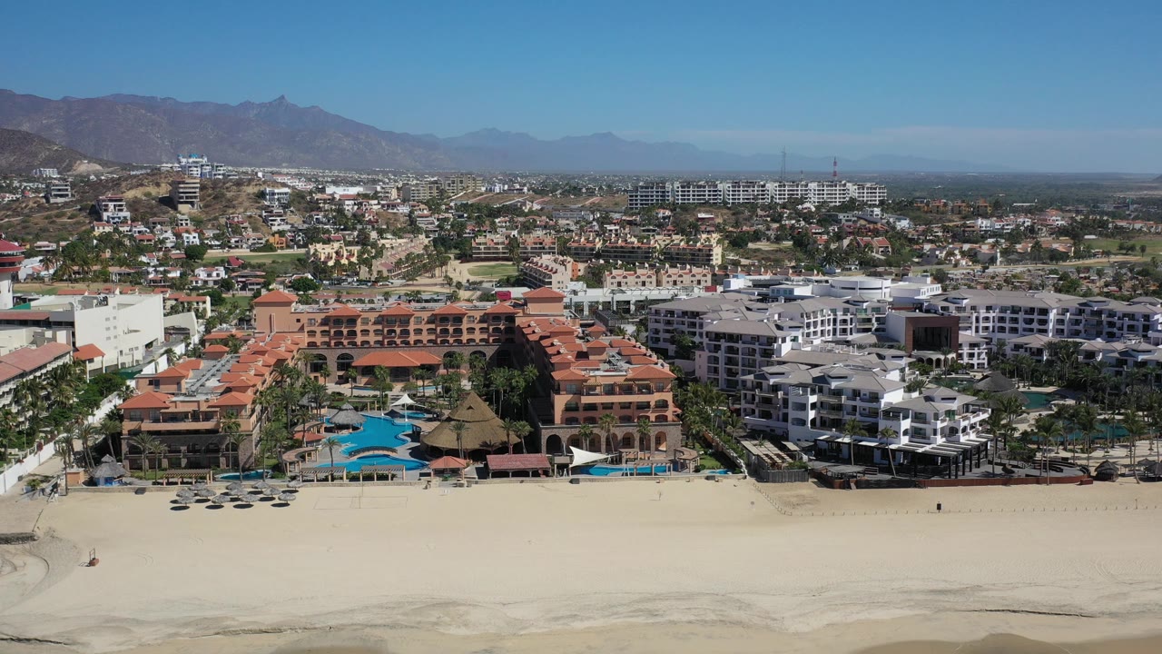 Aerial view in Los Cabos, Mexico showing beachfront resort buildings and homes above a wide pale beach, with distant mountains under clear daylight and tiny people on the sand.