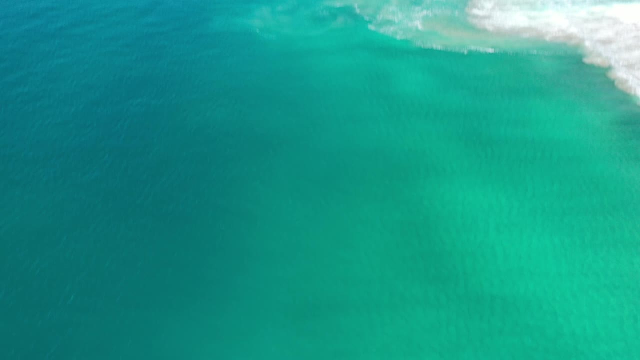 Aerial frame over green-blue Los Cabos water with a soft white wave band along the top edge and no visible people.