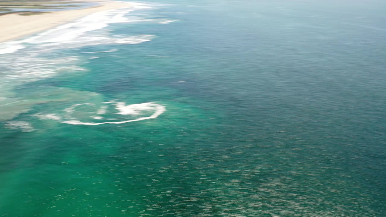 Aerial view of turquoise ocean with surf near a sandy beach.