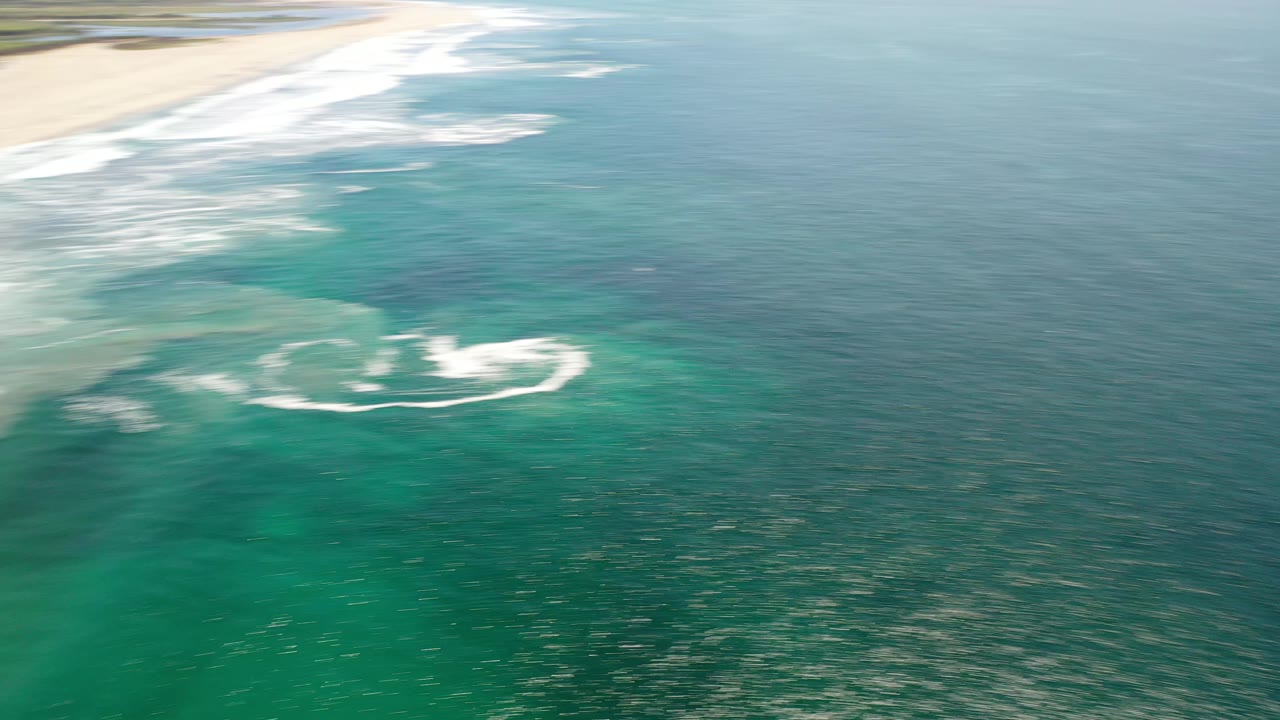 High aerial view in Los Cabos showing whitewater curling over green-blue ocean while the sandy shoreline remains near the top edge, with no visible people.