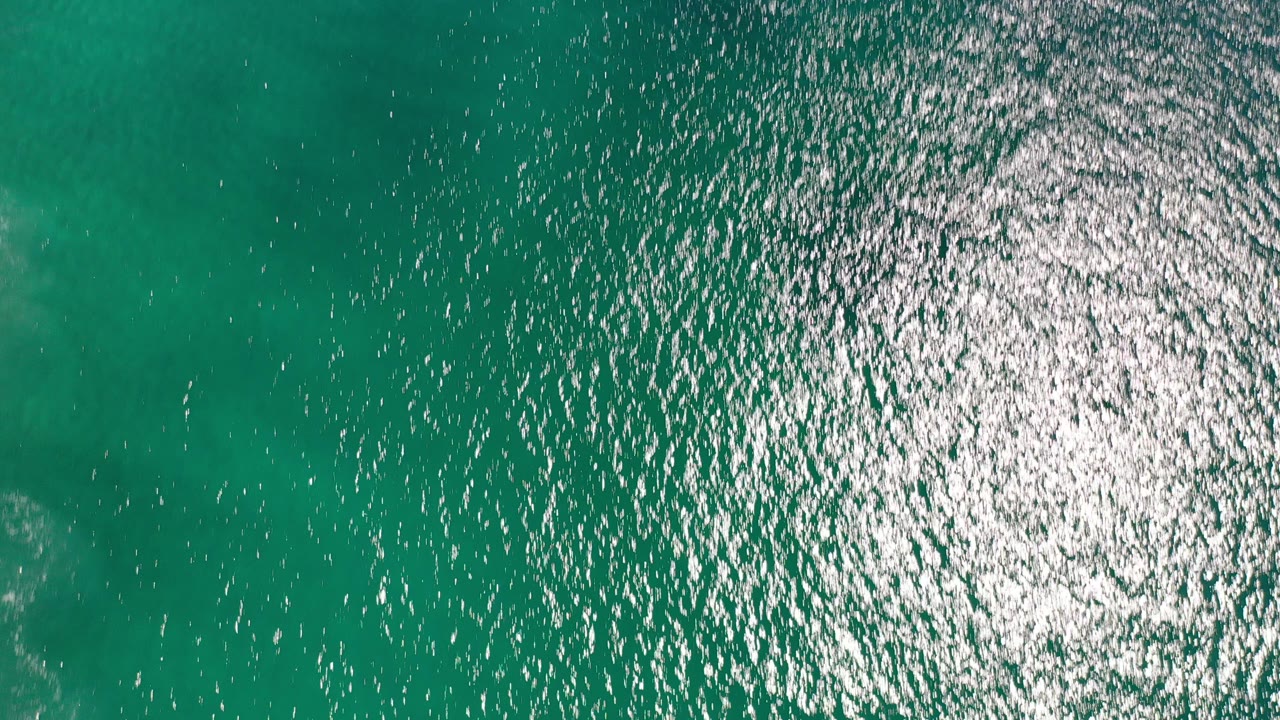 Overhead view of turquoise ocean water in Los Cabos, Mexico, with dense white sun sparkle near the upper center and no visible people.