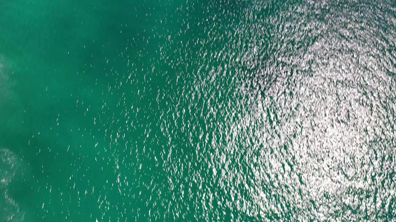 Aqua ocean water in Los Cabos, Mexico, fills the frame from above, with a broad glinting reflection across the middle and no visible people.