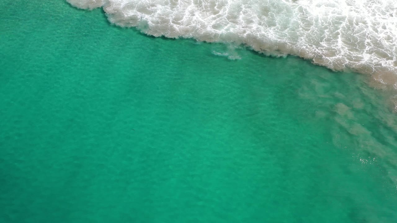 Overhead Los Cabos ocean frame with a thicker white surf band pushing across the top and translucent green water below, with no visible people.