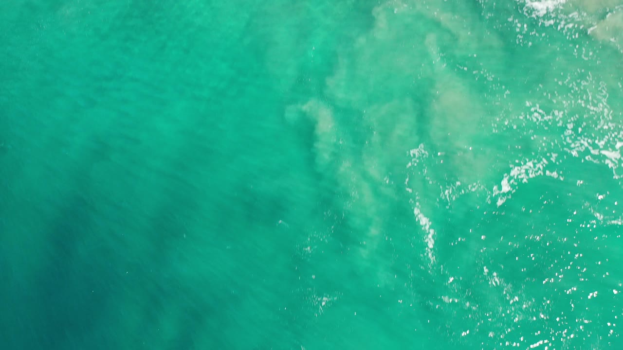 Overhead view of turquoise green ocean water in Los Cabos, Mexico, with pale foam patches, subtle reef tones below the surface, and no visible people.
