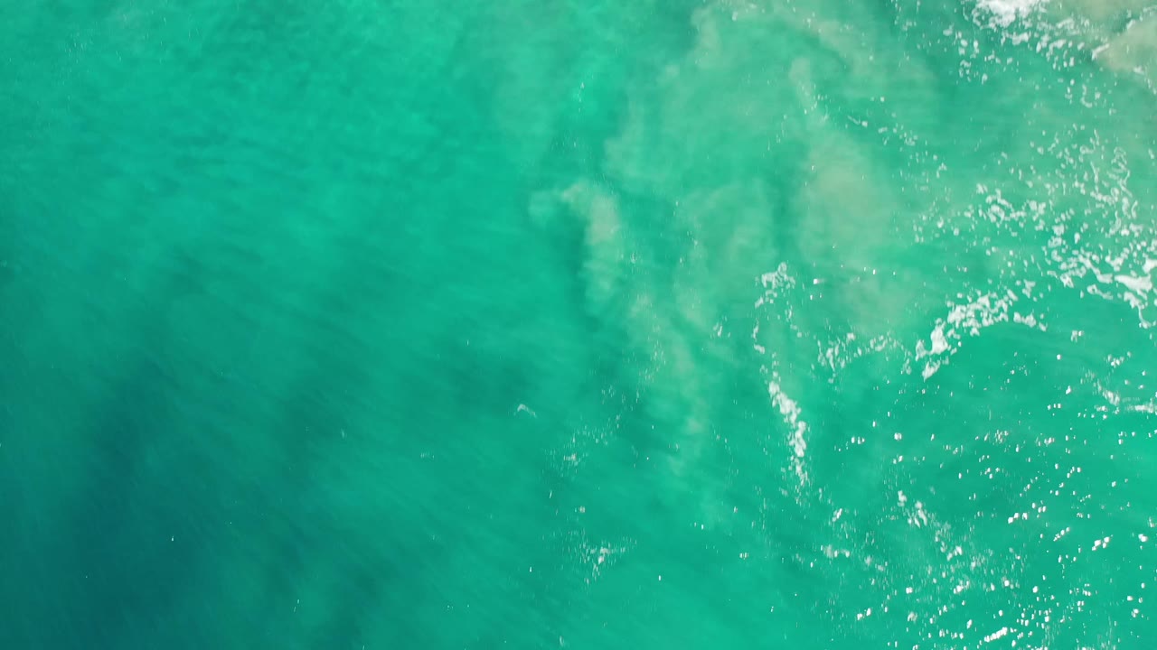 Aerial Los Cabos ocean surface filled with green water, scattered white foam, bright reflected sunlight, and no visible people.