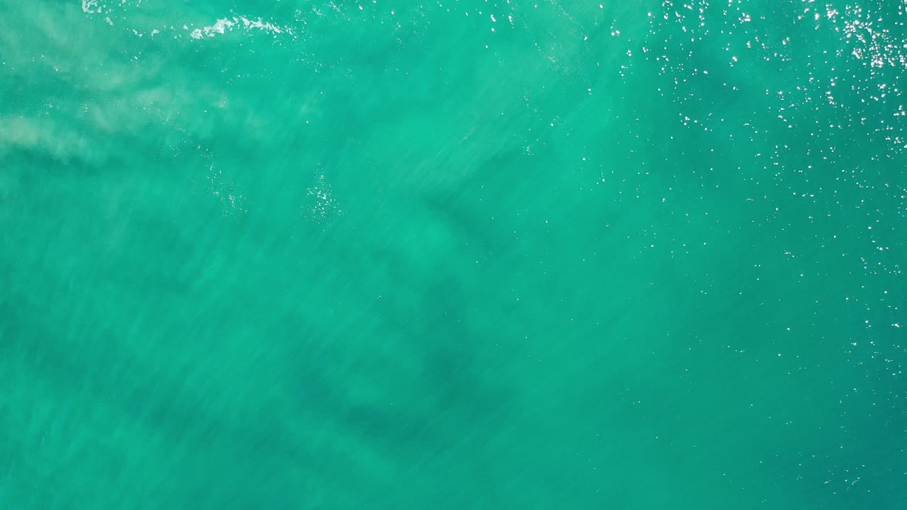 Top-down view of turquoise ocean in Los Cabos, Mexico, with muted sandy shapes under the water and no visible people.