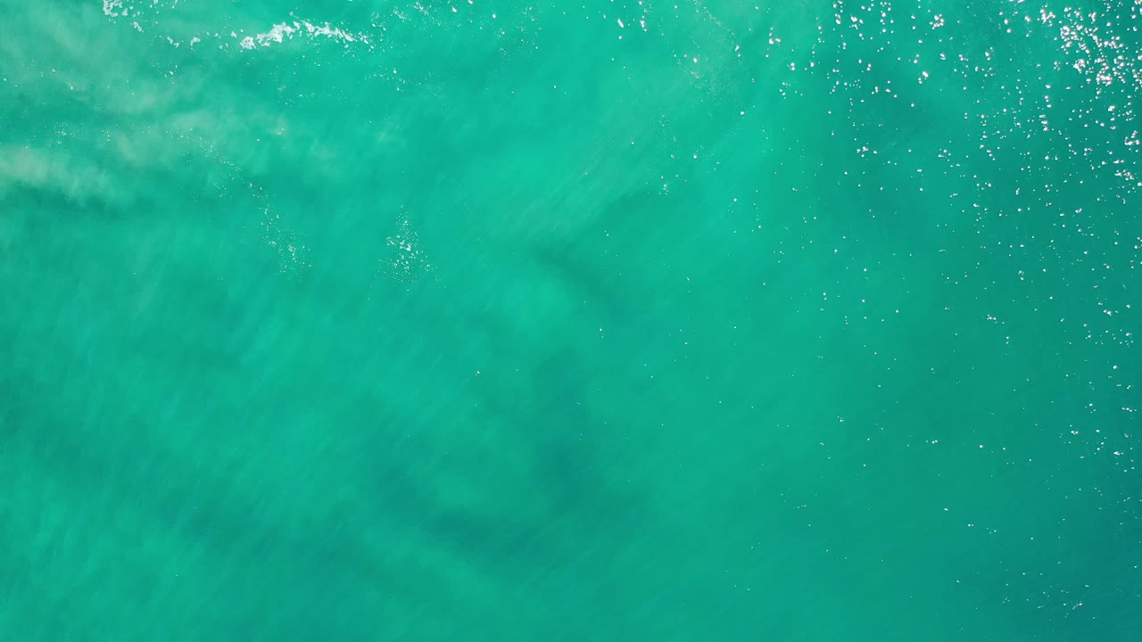 Overhead Los Cabos ocean frame with green-blue water, a small pale wave streak near the upper left, and no visible people.