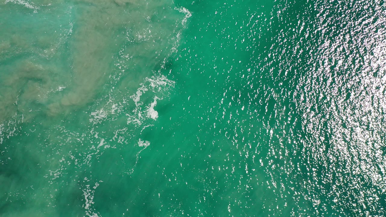Aerial view of blue-green ocean in Los Cabos with sun glitter across the right side, muted shallow patches beneath the water, and no visible people.