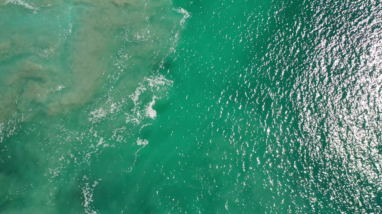 Aerial frame over Los Cabos water with a broad turquoise field, pale submerged reef or sand patches near the upper left, strong sun reflection on the right, and no visible people.