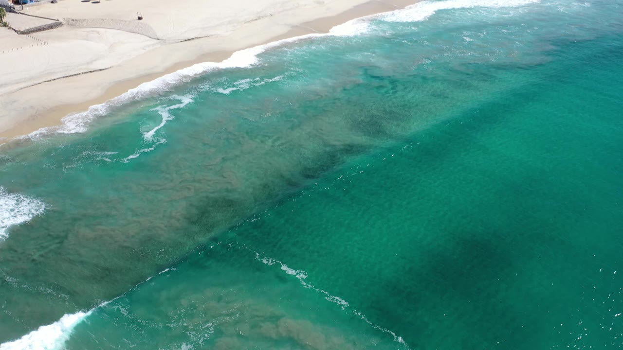 Aerial Los Cabos shoreline view with the beach still along the upper edge, blue-green water covering most of the frame, thin white wave lines, and no visible people.