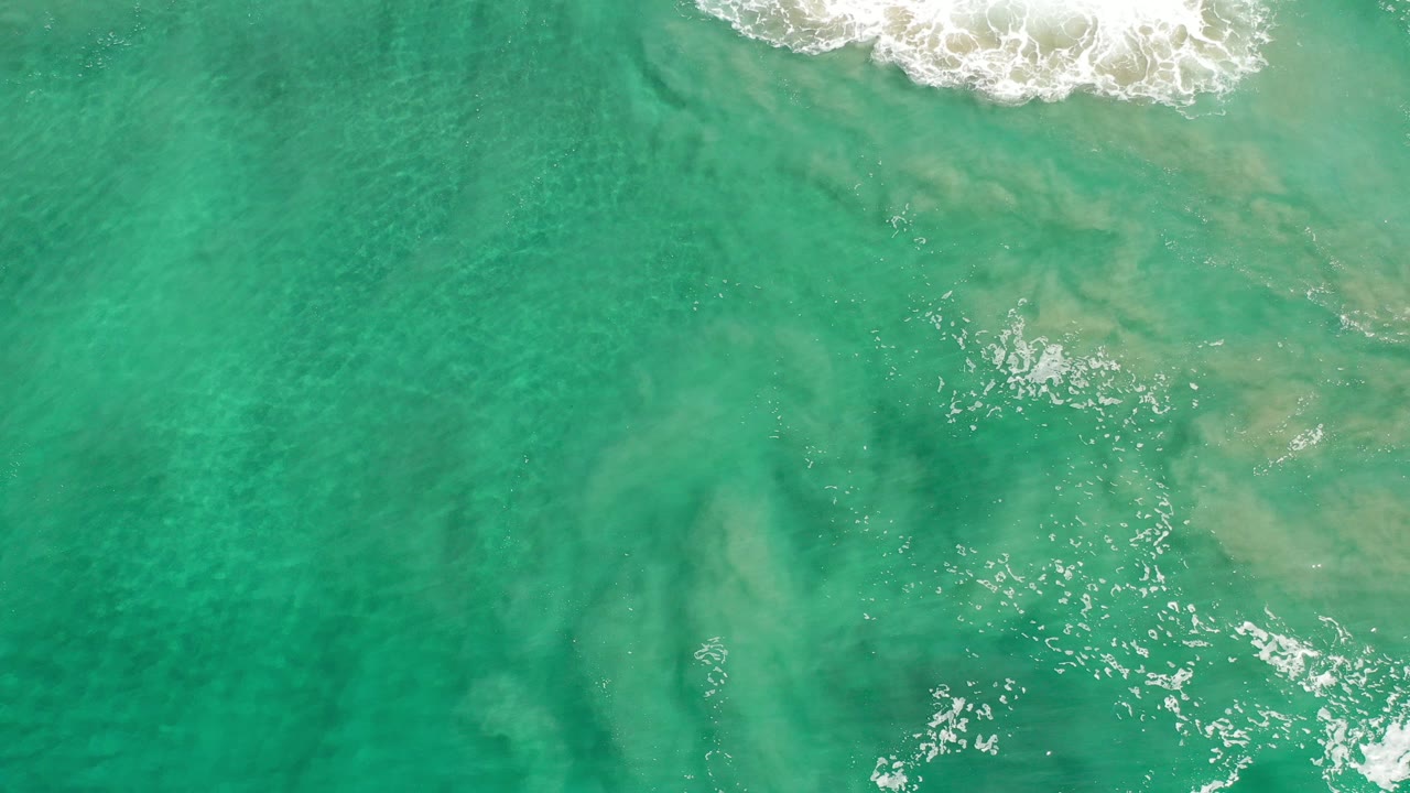 Overhead view of turquoise water in Los Cabos, Mexico, with white surf along the top edge and pale underwater patterns below, no visible people.