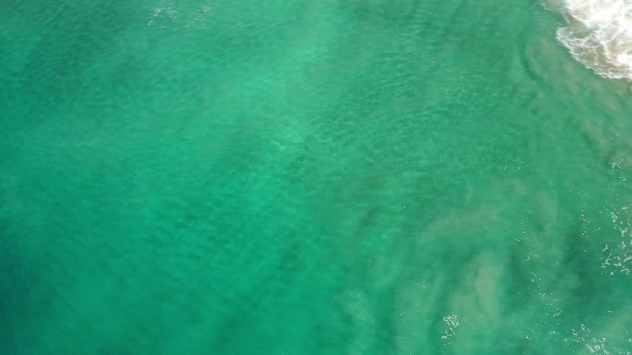 Top-down Los Cabos ocean view with clear green water, subtle underwater shapes, and white wave foam crossing the upper edge, no visible people.