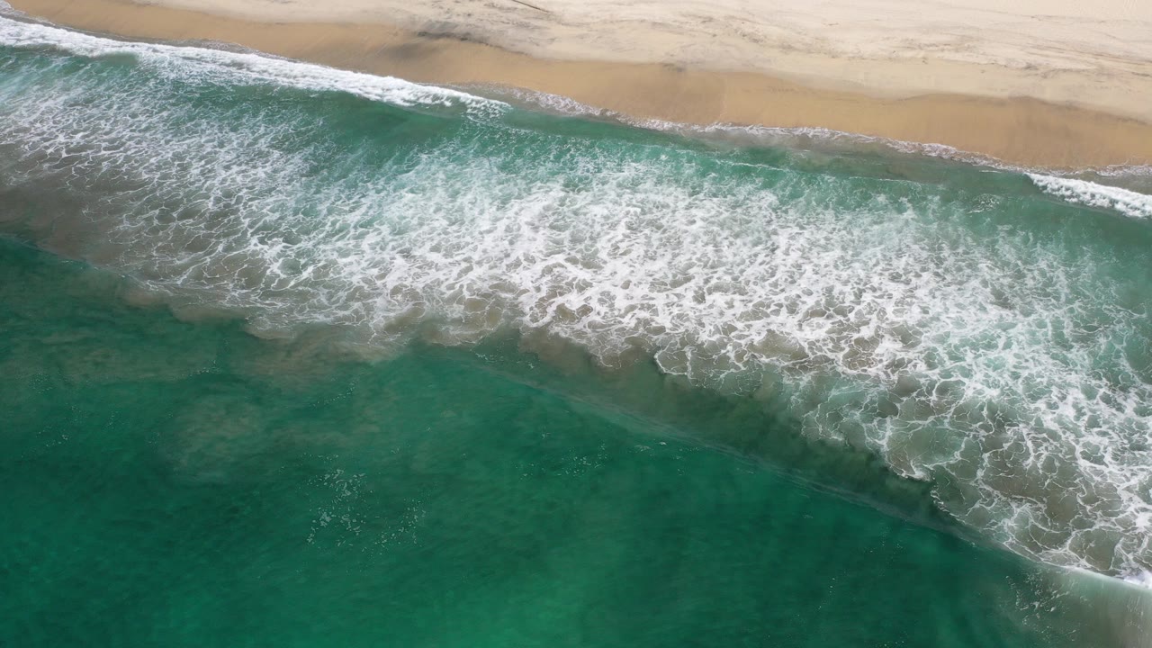 Overhead view of white surf breaking diagonally along a sandy Los Cabos shoreline, with green-blue water below and no visible people.