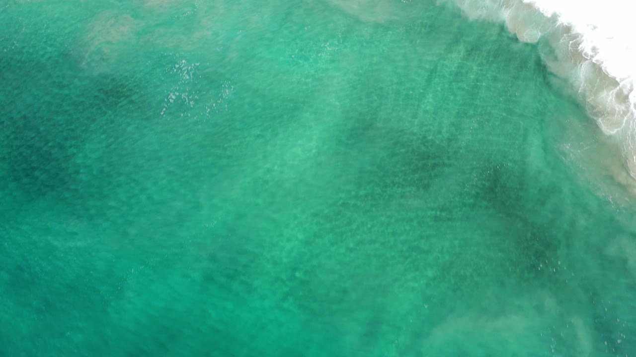 Overhead view of turquoise Los Cabos ocean water with white surf crossing the upper right edge, darker underwater patches, and no visible people.