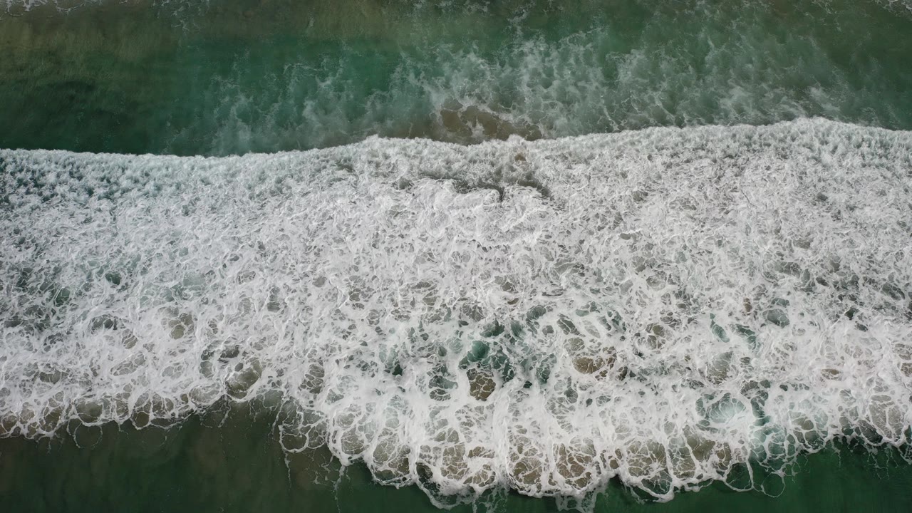 Overhead view in Los Cabos, Mexico, with dense white foam spreading across dark teal ocean water and no visible people.