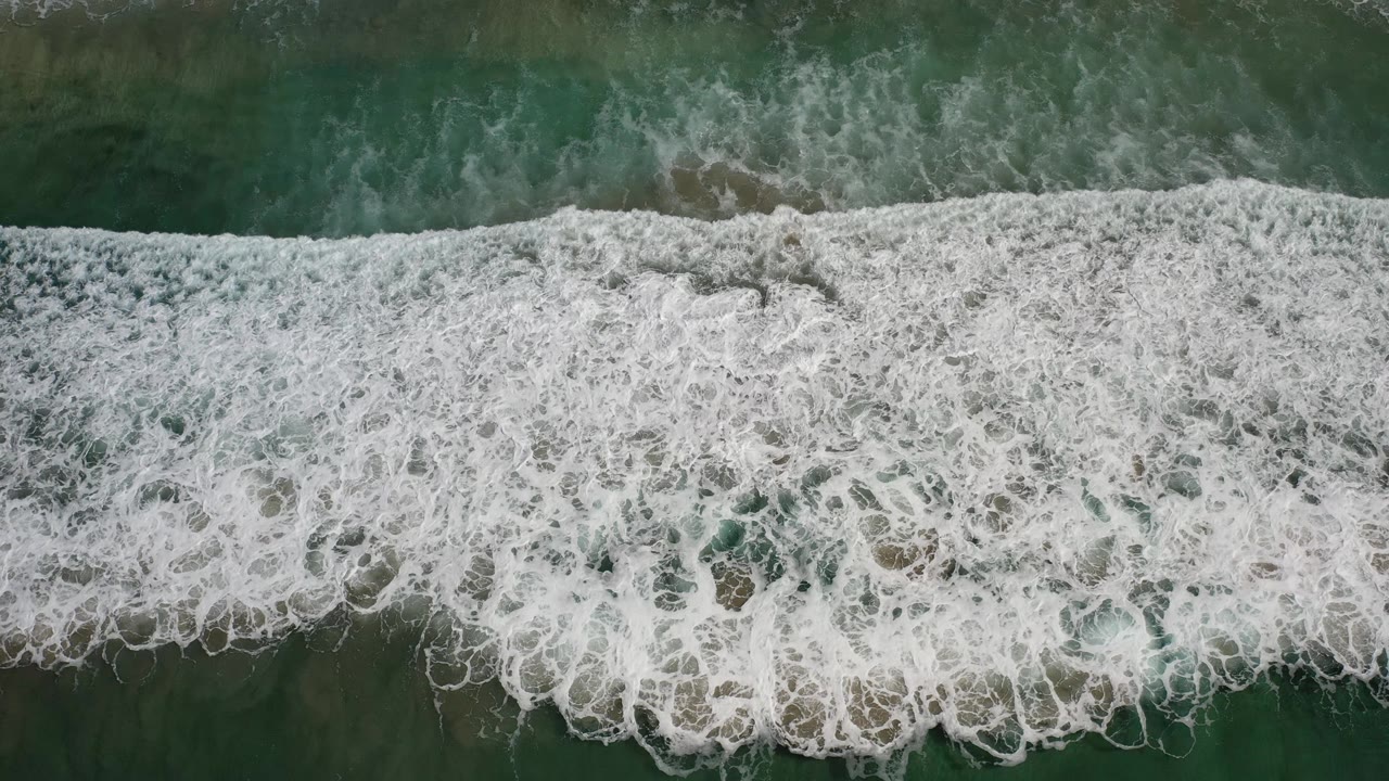 Aerial frame of Los Cabos surf where white foam covers most of the water surface, with green-teal patches visible beneath and no visible people.