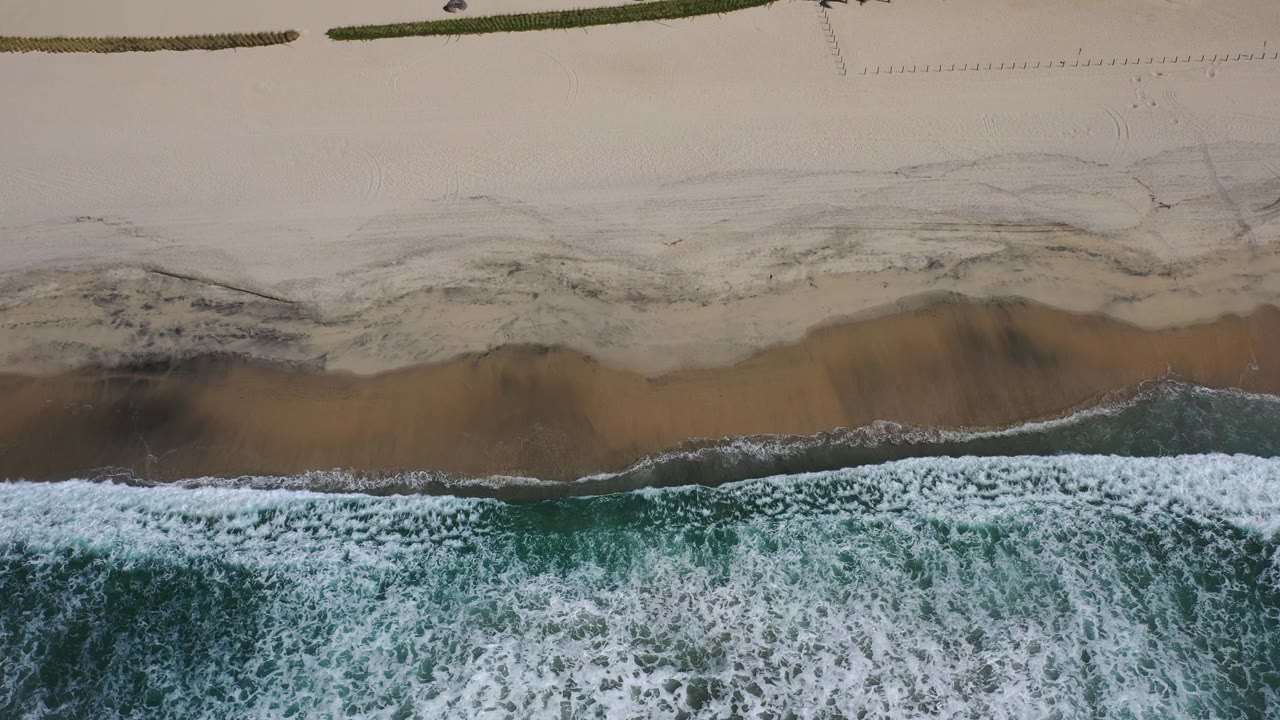 An overhead Los Cabos beach frame shows a broad band of white wave foam meeting brown wet sand and pale dry sand, with no visible people.