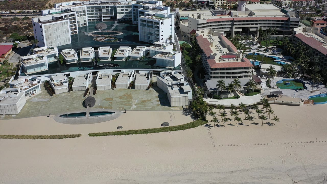 High oblique aerial frame over Los Cabos, Mexico showing white beachfront hotel buildings, palm rows, water features, wide sand, hillside development, and no visible people.