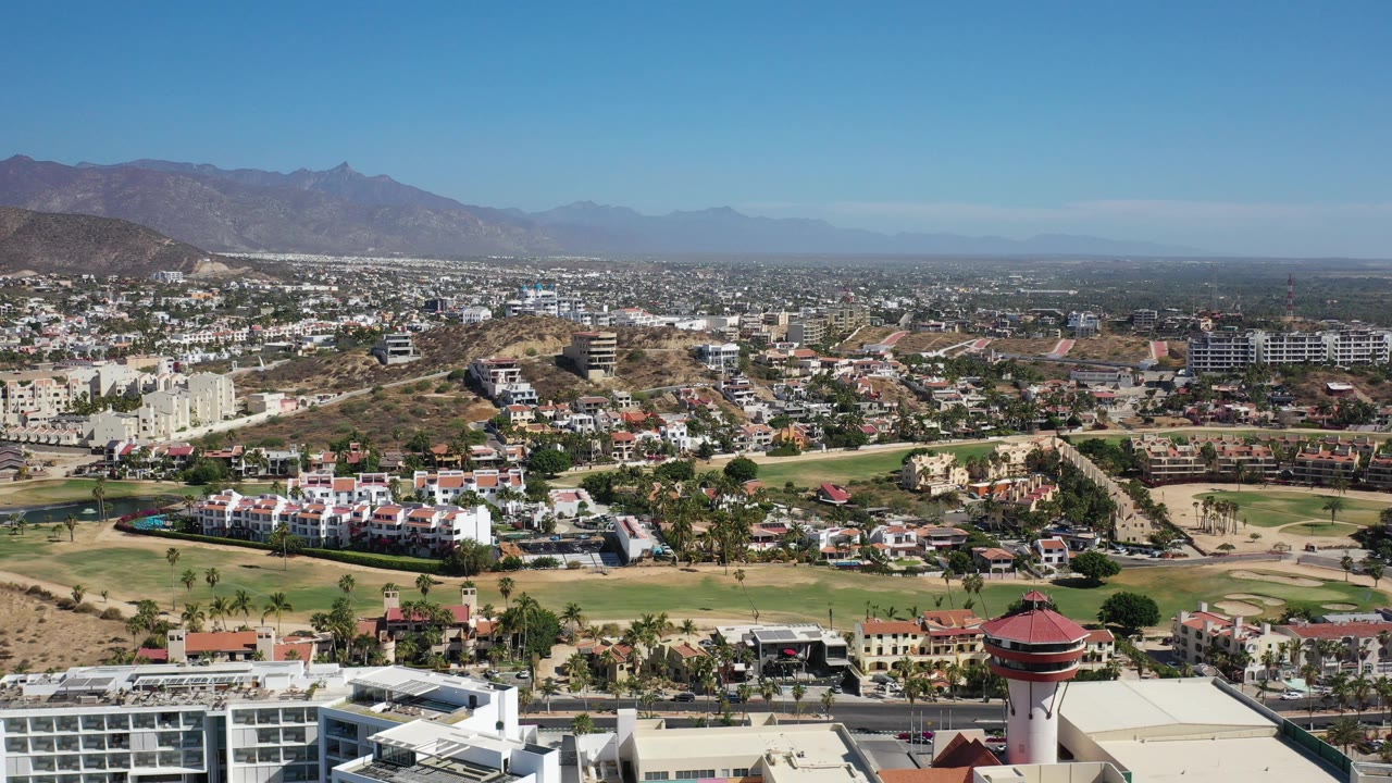 Second aerial frame in Los Cabos, Mexico, with the landscaped green area and white buildings more centered, dry hills behind them, distant city blocks, and no visible people.