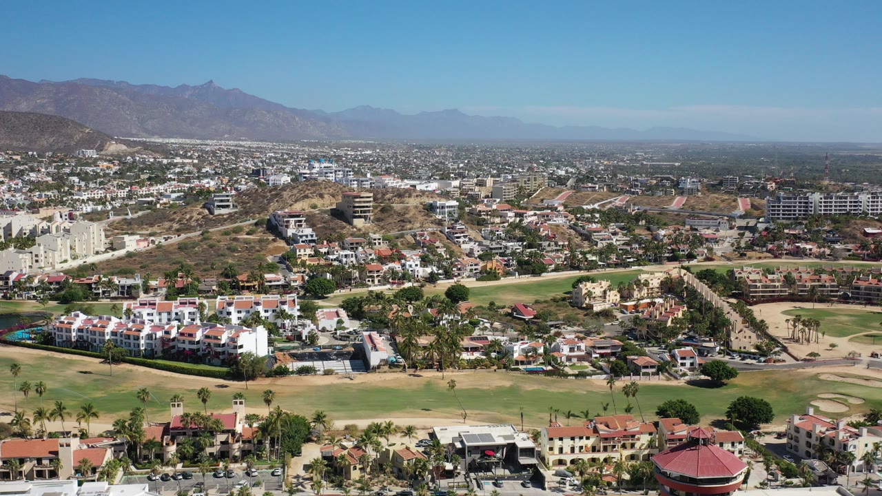 Wide aerial view of Los Cabos, Mexico, showing red-roofed residences along the bottom edge, green golf-course fairways, dry hills, distant town blocks, mountains, and no visible people.