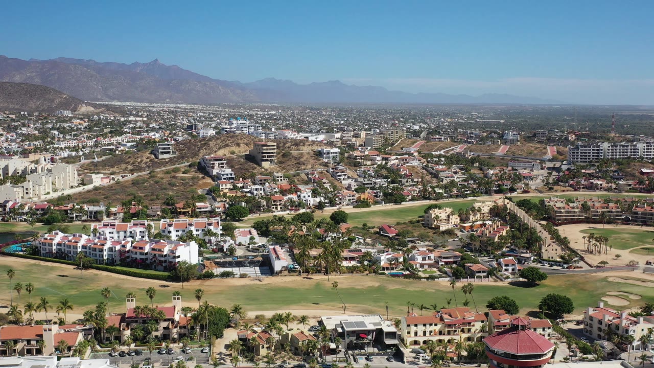 Elevated Los Cabos aerial with a road and rooftops in the foreground, fairways across the middle, hillside residences and larger white buildings beyond, distant mountains, and no visible people.