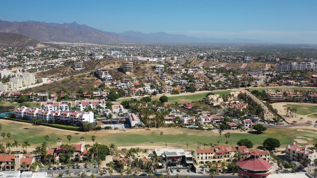 Drone view across a Los Cabos neighborhood with fairways, homes, hills, and open sky.