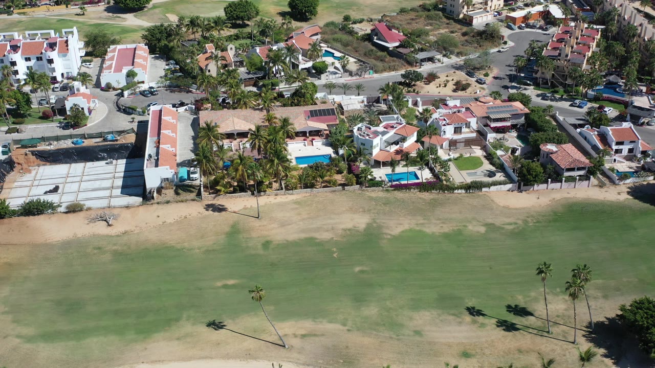 Los Cabos drone view with the golf course covering the lower frame, a long white building, red tile roofs, blue pools, palms, roads, and no visible people.