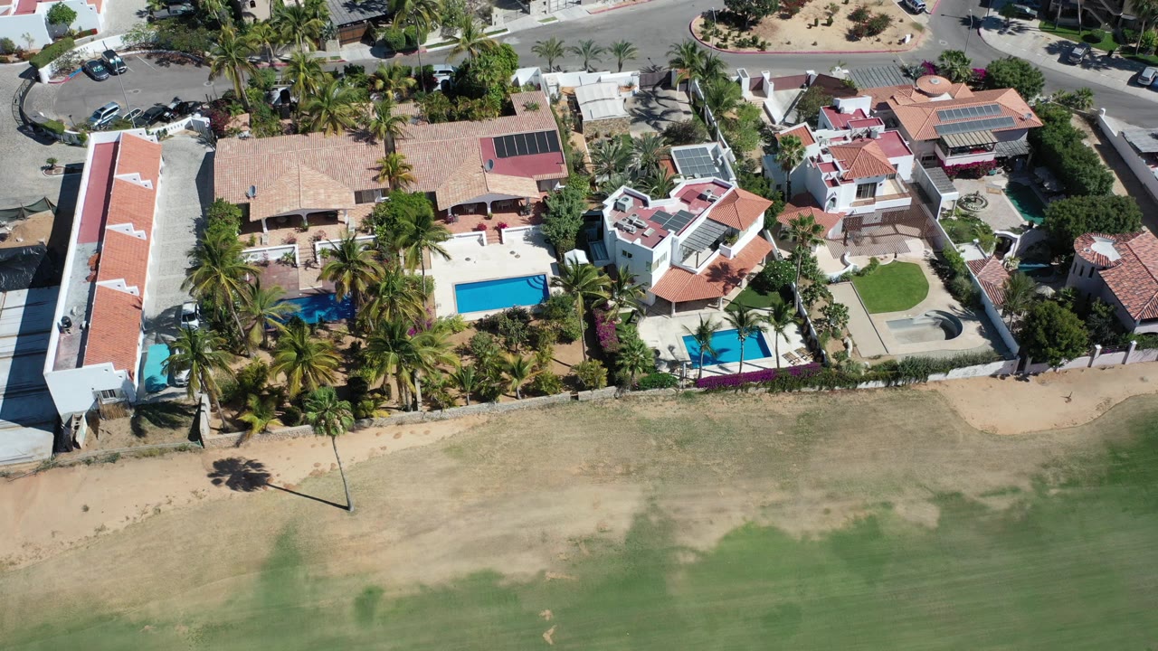 Overhead view of tiled-roof buildings, pools, palms, sandy ground, and a street.