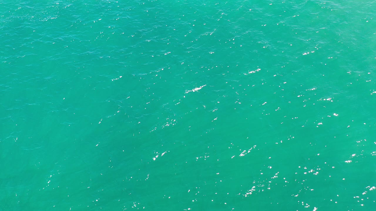 Turquoise ocean water in Los Cabos, Mexico fills the frame at the start of the clip, with scattered white sun reflections and no visible people.