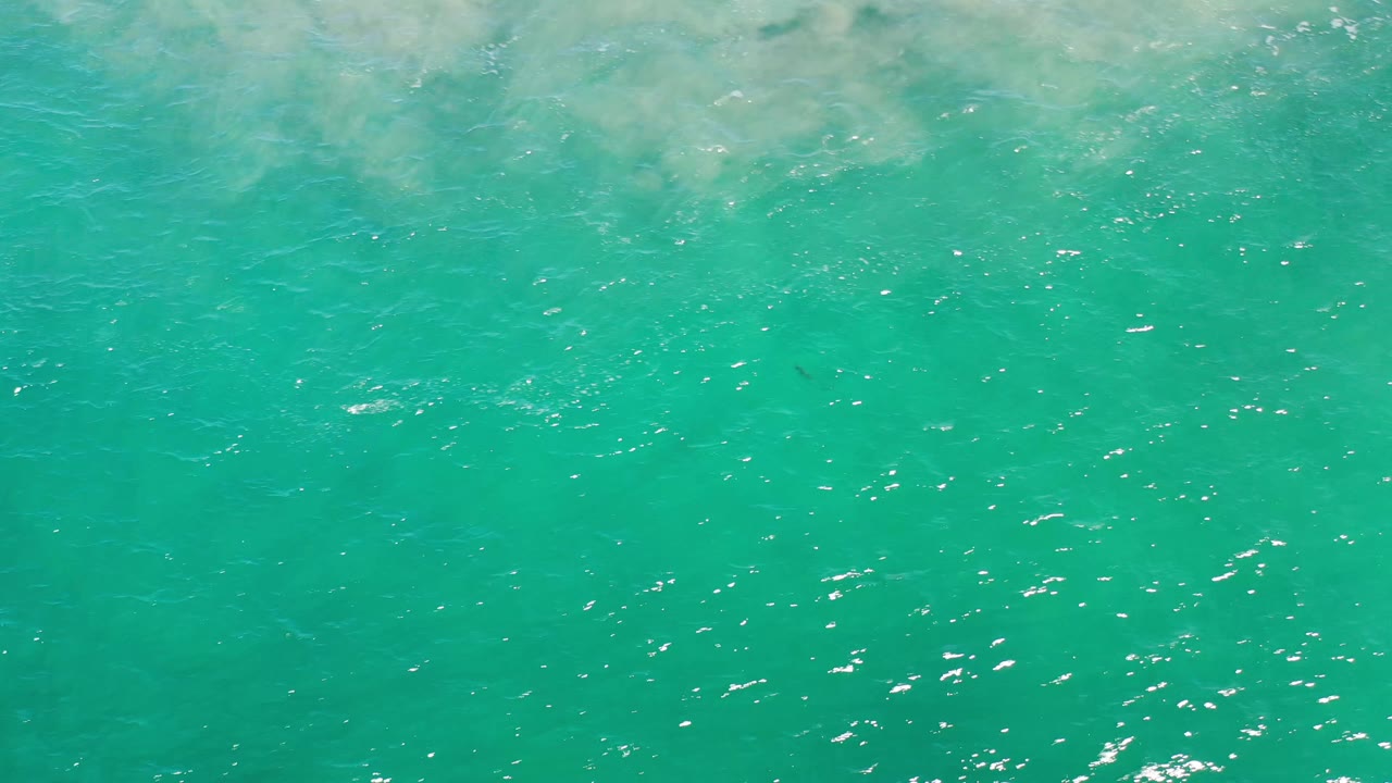 Overhead Los Cabos water frame showing teal sea, scattered sun reflections, and lighter sandy color near the upper edge, with no visible people.