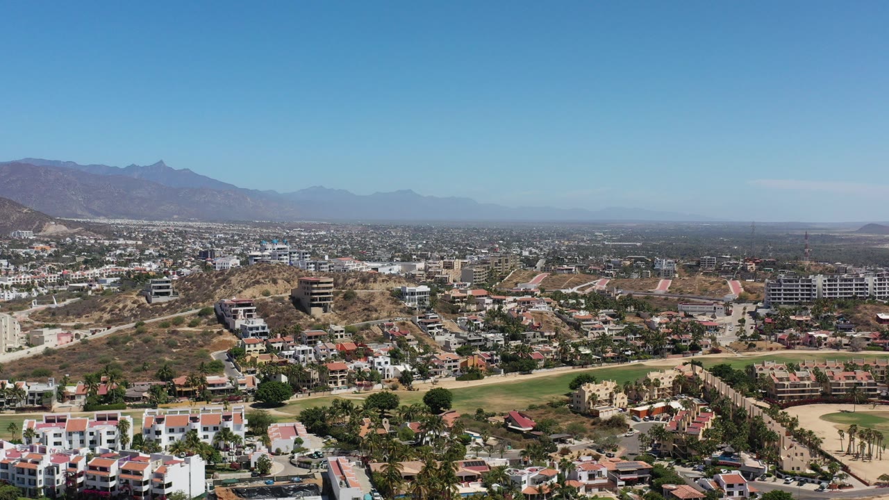 Wide aerial view of hillside residences, greenery, and distant mountains under blue sky.