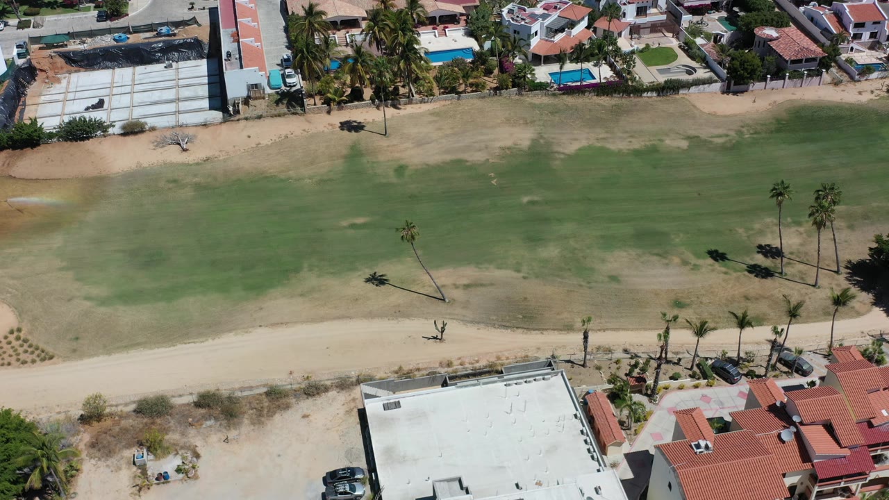 High aerial view over Los Cabos homes, palms, a sandy road, and a broad patchy green fairway, with no visible people.