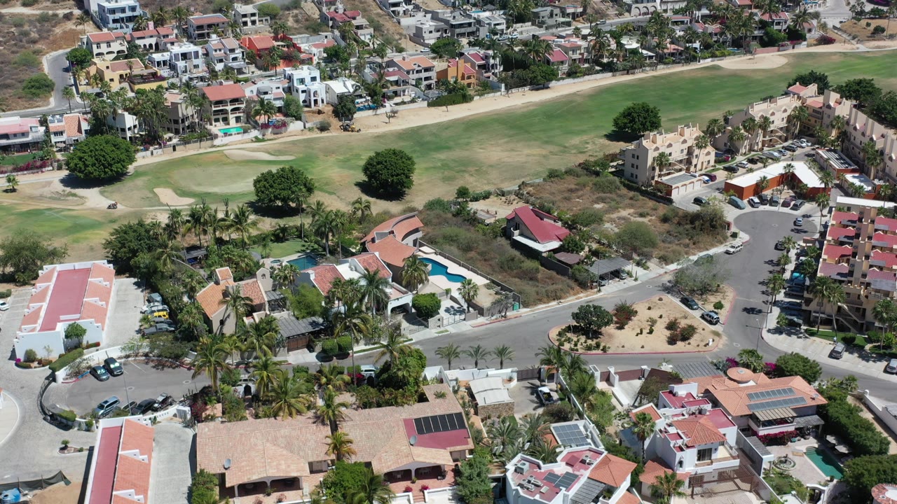 Aerial view over Los Cabos hillside homes, apartment buildings, curved streets, palms, pools, and golf-course grass with no visible people.