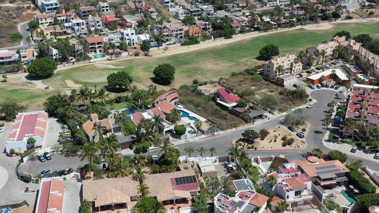Drone view across a Los Cabos residential hillside, with red-roof homes, apartment blocks, palm trees, parked cars, green fairway, and no visible people.
