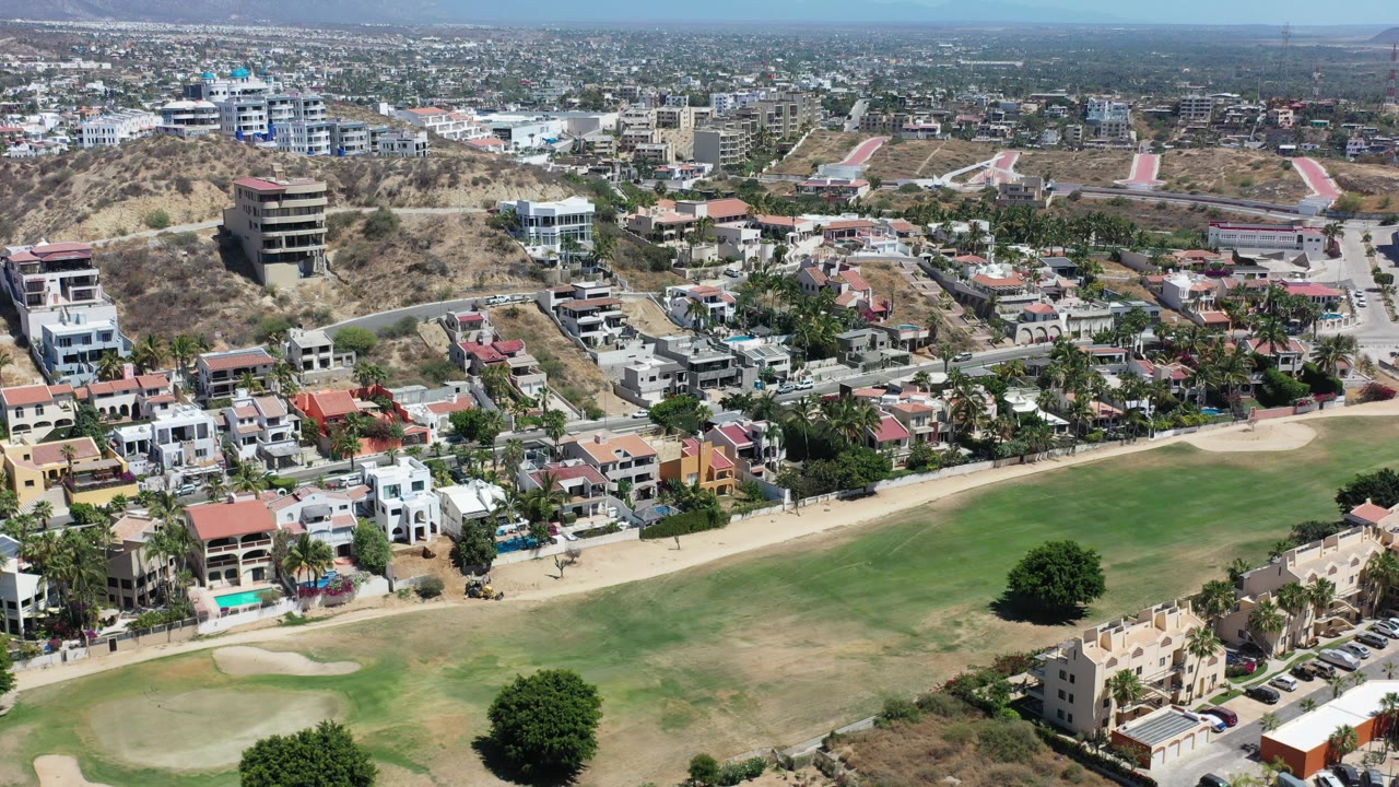 Los Cabos aerial frame with hillside houses, a green fairway in the foreground, dry slopes, spread-out city blocks, distant mountains, and no visible people.