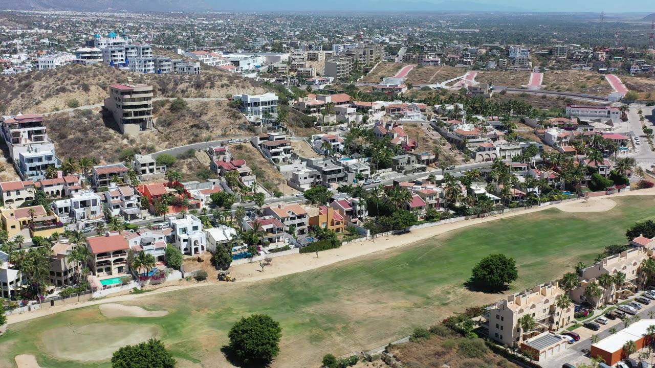 Los Cabos aerial view centered on a dry hill with homes below, palm trees, roadways, low-rise buildings, distant urban sprawl, mountains, and no visible people.