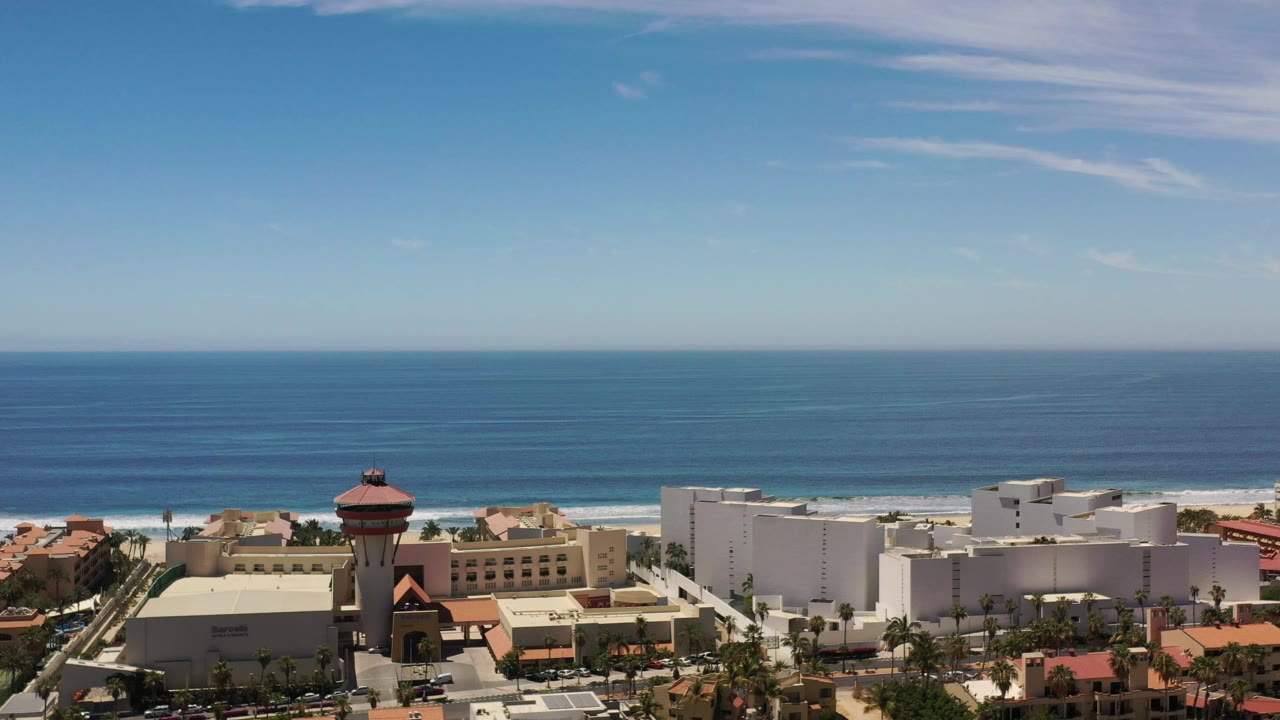 High aerial frame in Los Cabos showing white resort structures in the foreground, a narrow beach band, low surf, open ocean, and no visible people.