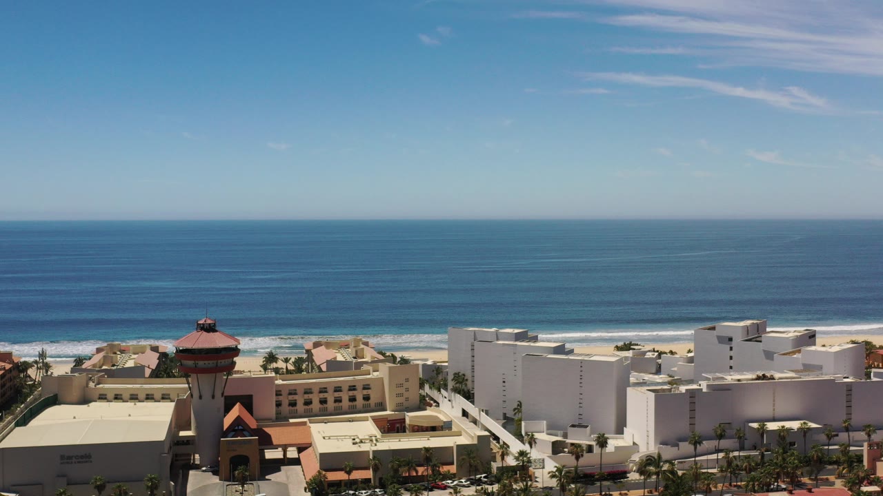 Aerial view over Los Cabos, Mexico, with low-rise buildings in the foreground, a sandy beach and blue ocean beyond, and no visible people.