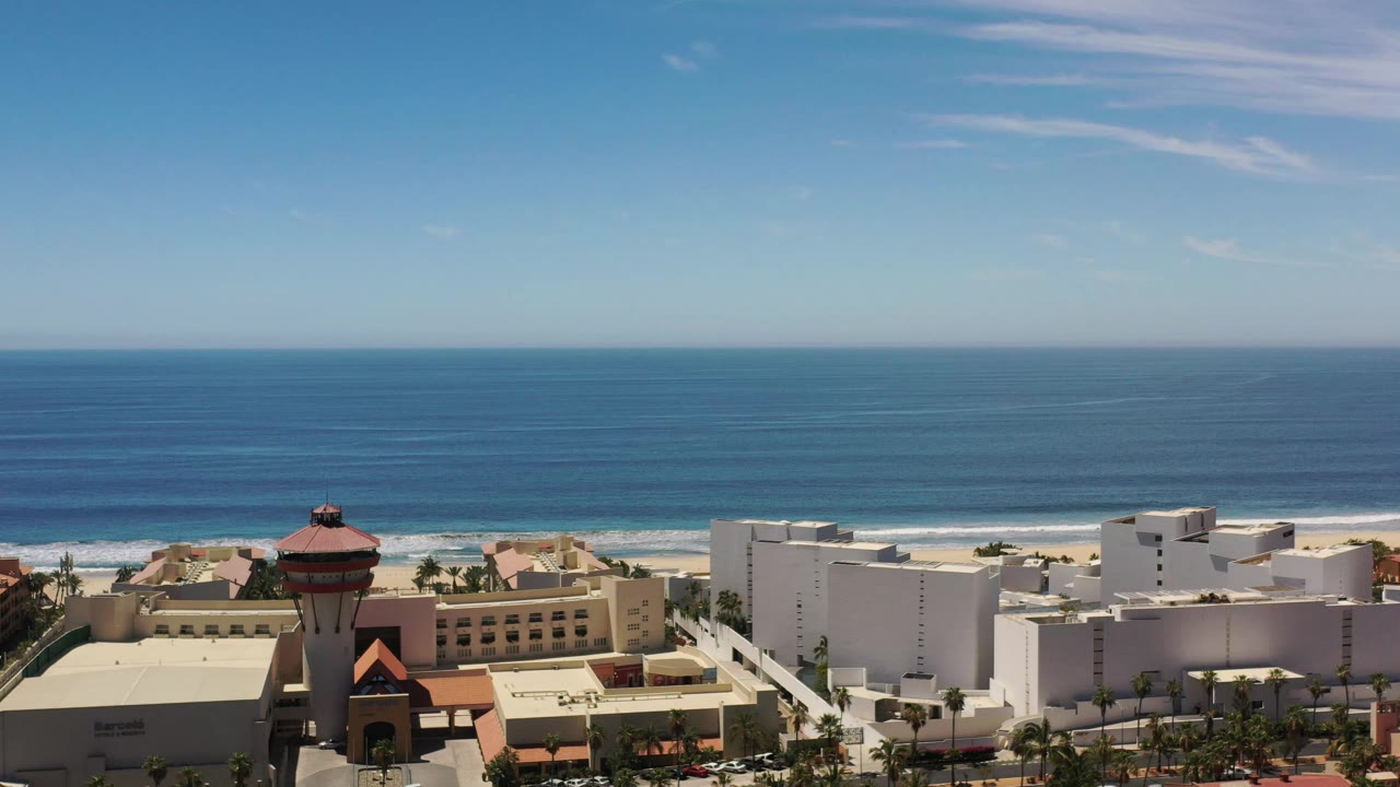 Los Cabos aerial frame with a red-roofed tower near the lower center, white buildings to the right, beach behind them, and no visible people.