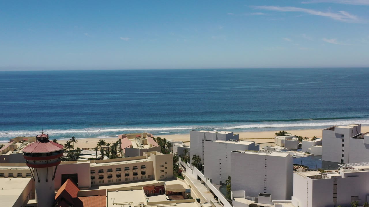 Elevated Los Cabos view with deep blue ocean beyond a sandy beach and white waterfront buildings in the foreground, no visible people.
