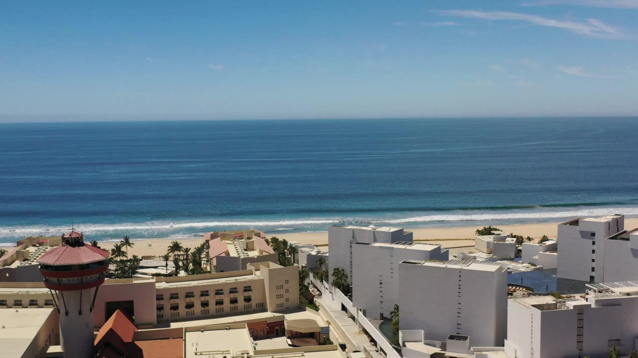 Los Cabos shoreline with a broad ocean horizon, breaking surf, palm trees, and blocky white coastal buildings, no visible people.