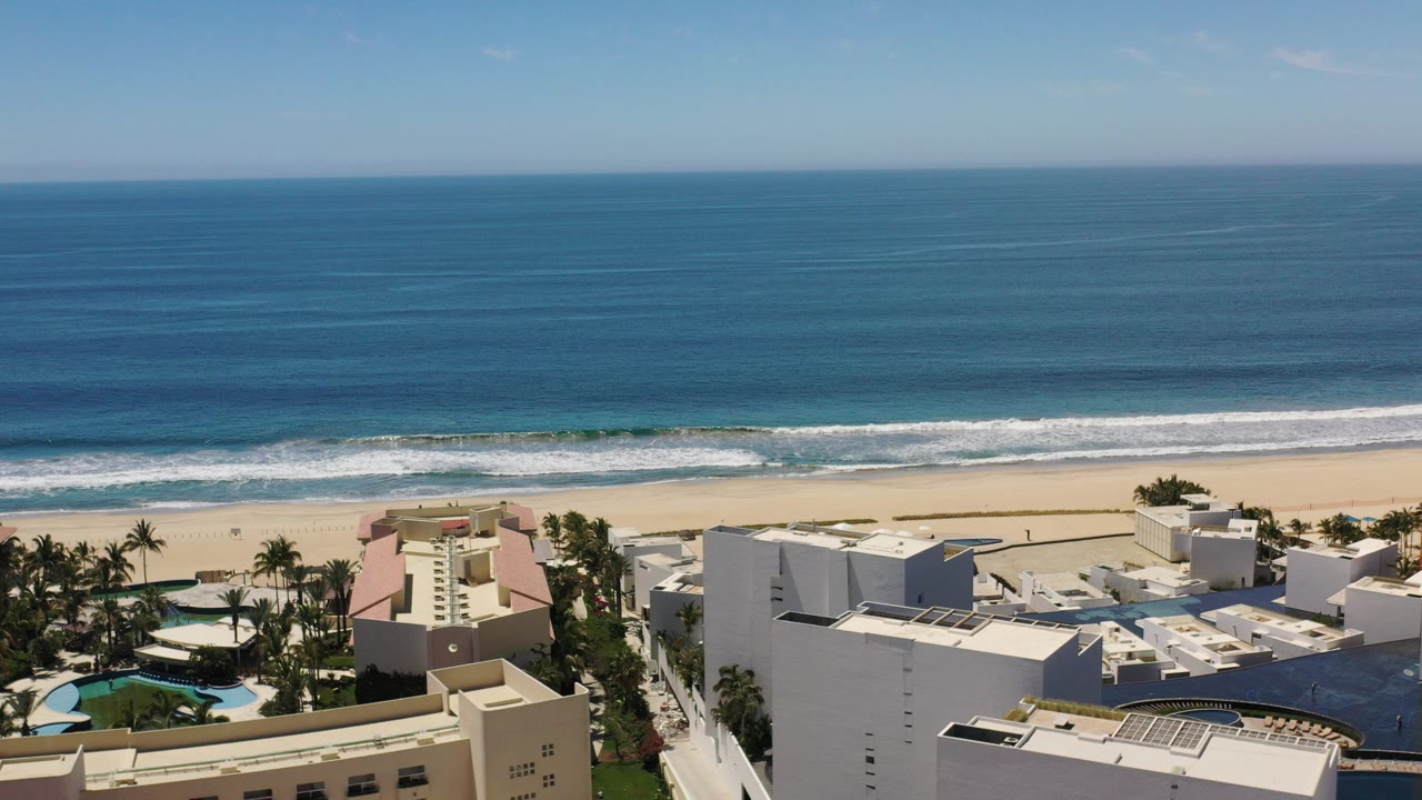 Opening aerial frame over Los Cabos, Mexico, with blue ocean, a band of white surf, wide sand, beachfront rooftops, palms, and no clearly visible people.