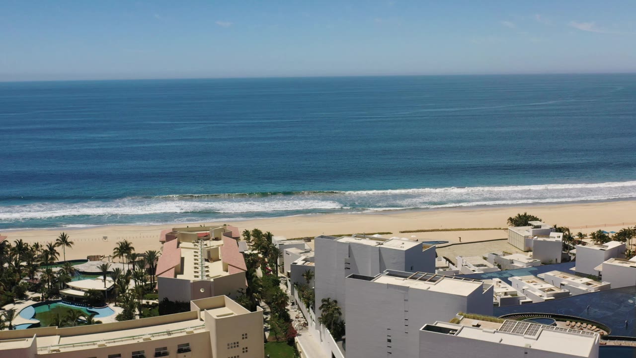 Aerial Los Cabos beach view with the surf line slightly shifted, pale sand below the waves, white rooftops, palms, pool areas, and no clearly visible people.
