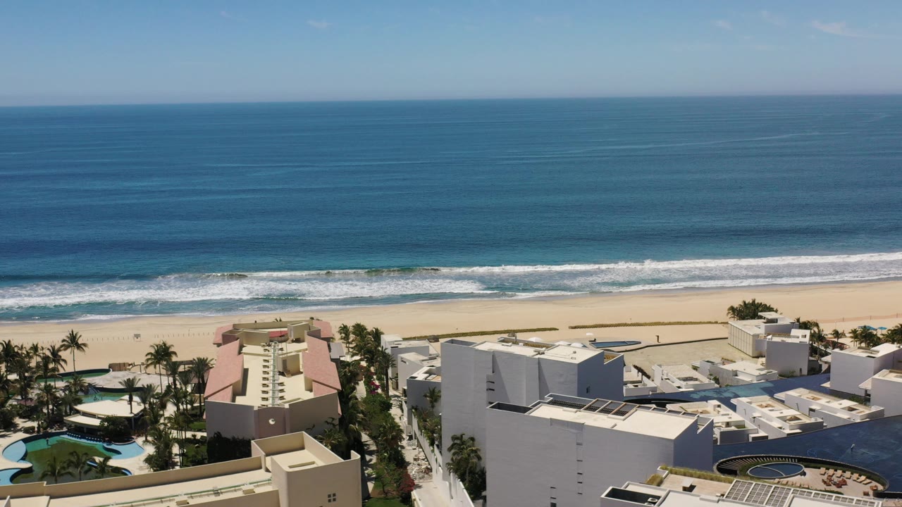 Ocean and beach with low buildings and palms along the shore.