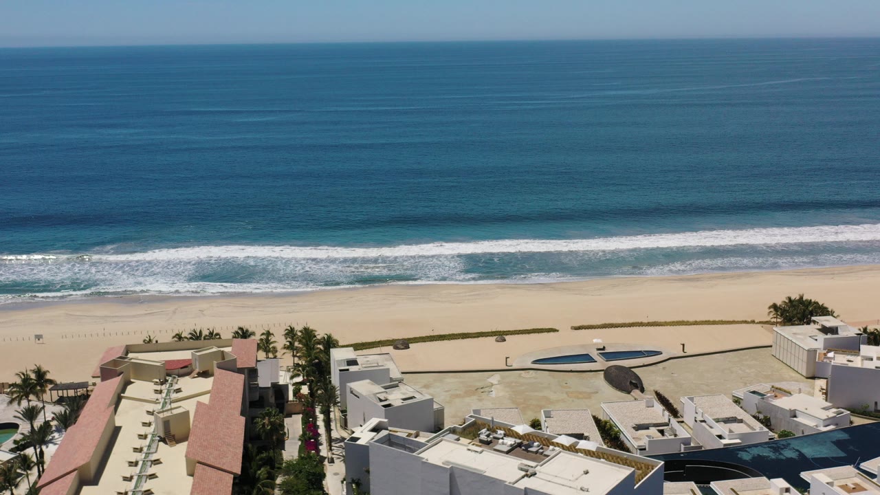 Aerial view in Los Cabos, Mexico, with teal ocean beyond white surf, pale sand, palm trees, red rooftops, pools, and no visible people.